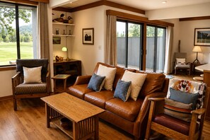 A cozy living room with a leather sofa, wooden coffee table, and armchairs, featuring large windows and a view of a green landscape.