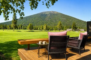 A wooden deck with wicker chairs and a table, overlooking a green field and a mountain.