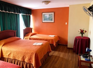 A hotel room with two beds, a small table with a lamp, a fan, and a painting on the wall.