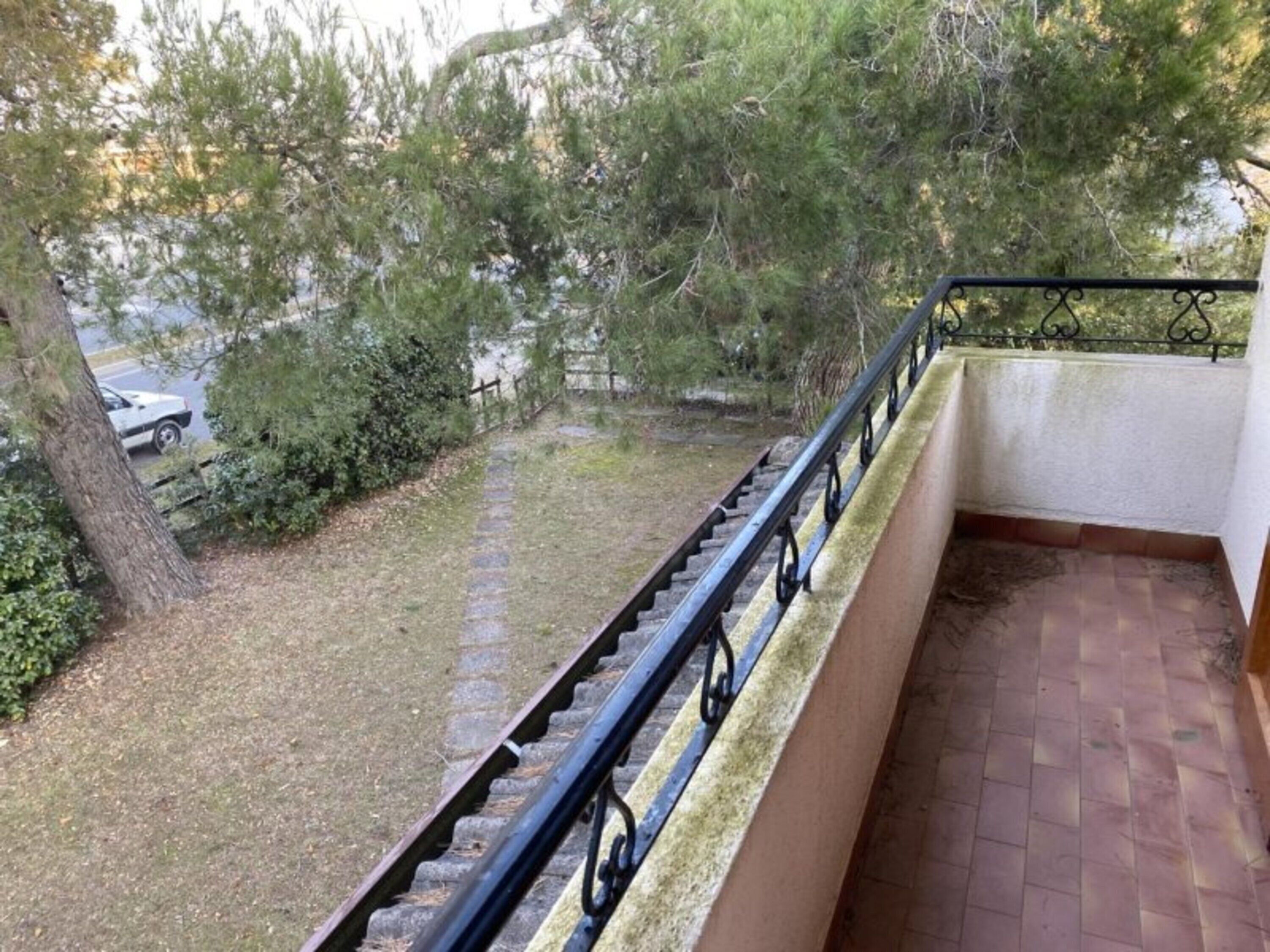 Un balcone con ringhiera in metallo, pavimento in pietra e vista su una zona verde alberata con un'auto parcheggiata.