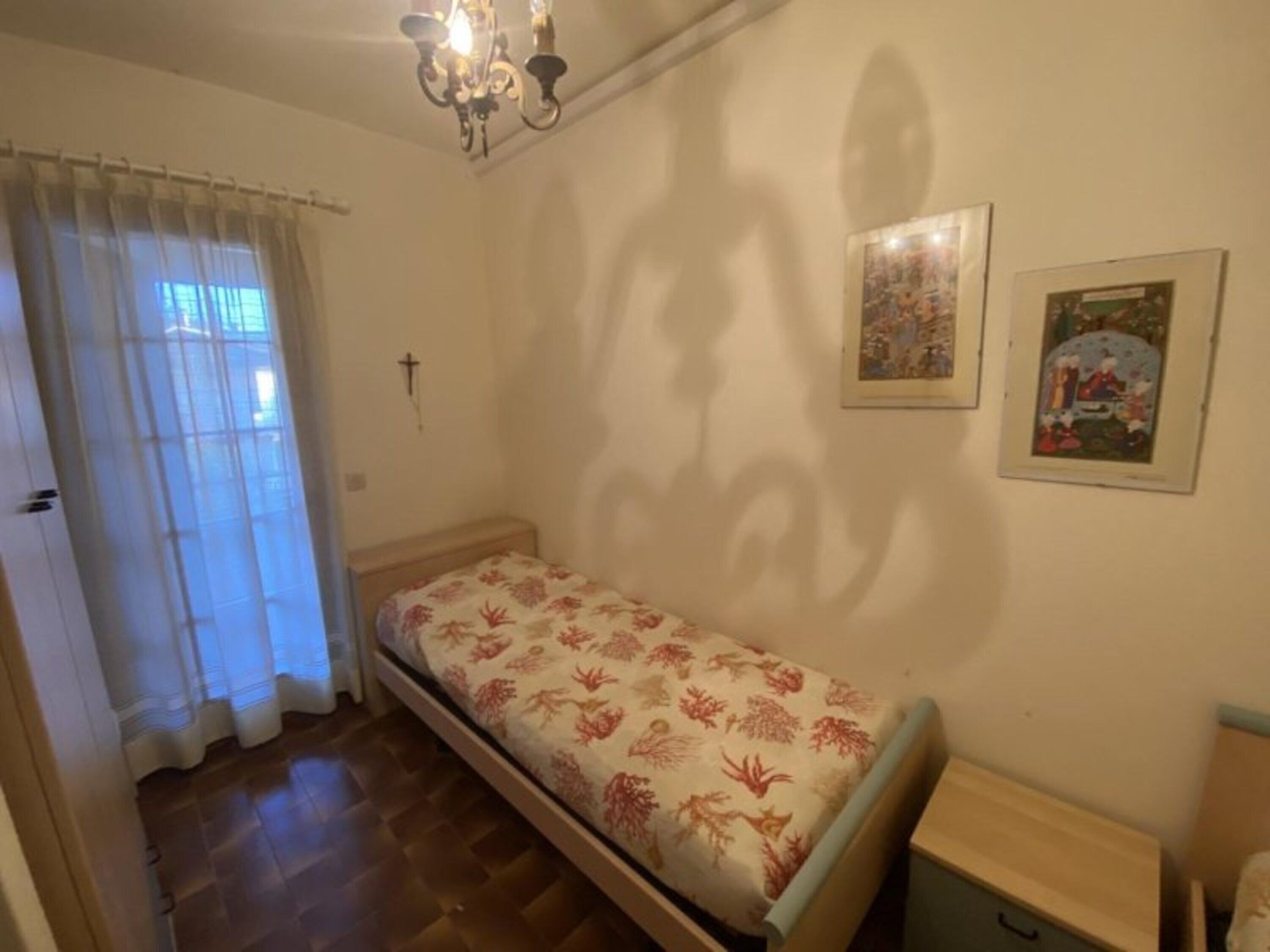A small bedroom with a single bed, a wooden floor, a chandelier, a cross on the wall, and two framed pictures.