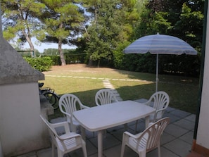 Eine Terrasse im Freien mit Tisch und Stühlen, einem gestreiften Sonnenschirm und Blick auf einen Rasen mit Bäumen.