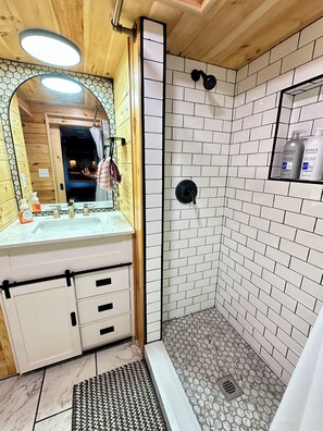 A bathroom with a wooden vanity, a shower with white tiles, and a mirror with a hexagonal tile backsplash.