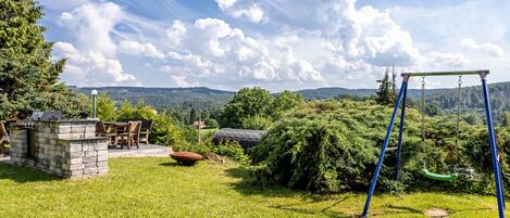 A grassy outdoor area with a stone barbecue station, a swing set, and a view of distant hills.