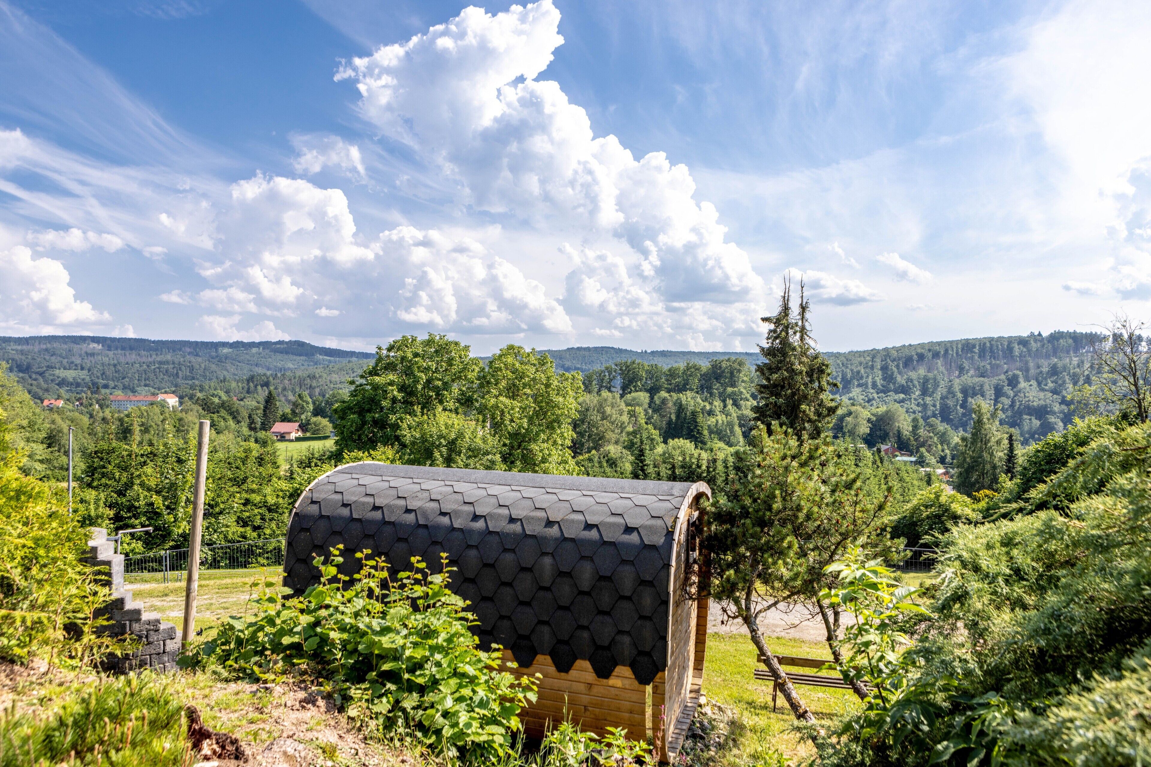 A small, wooden cabin with a hexagonal roof, surrounded by lush greenery and a mountainous landscape.