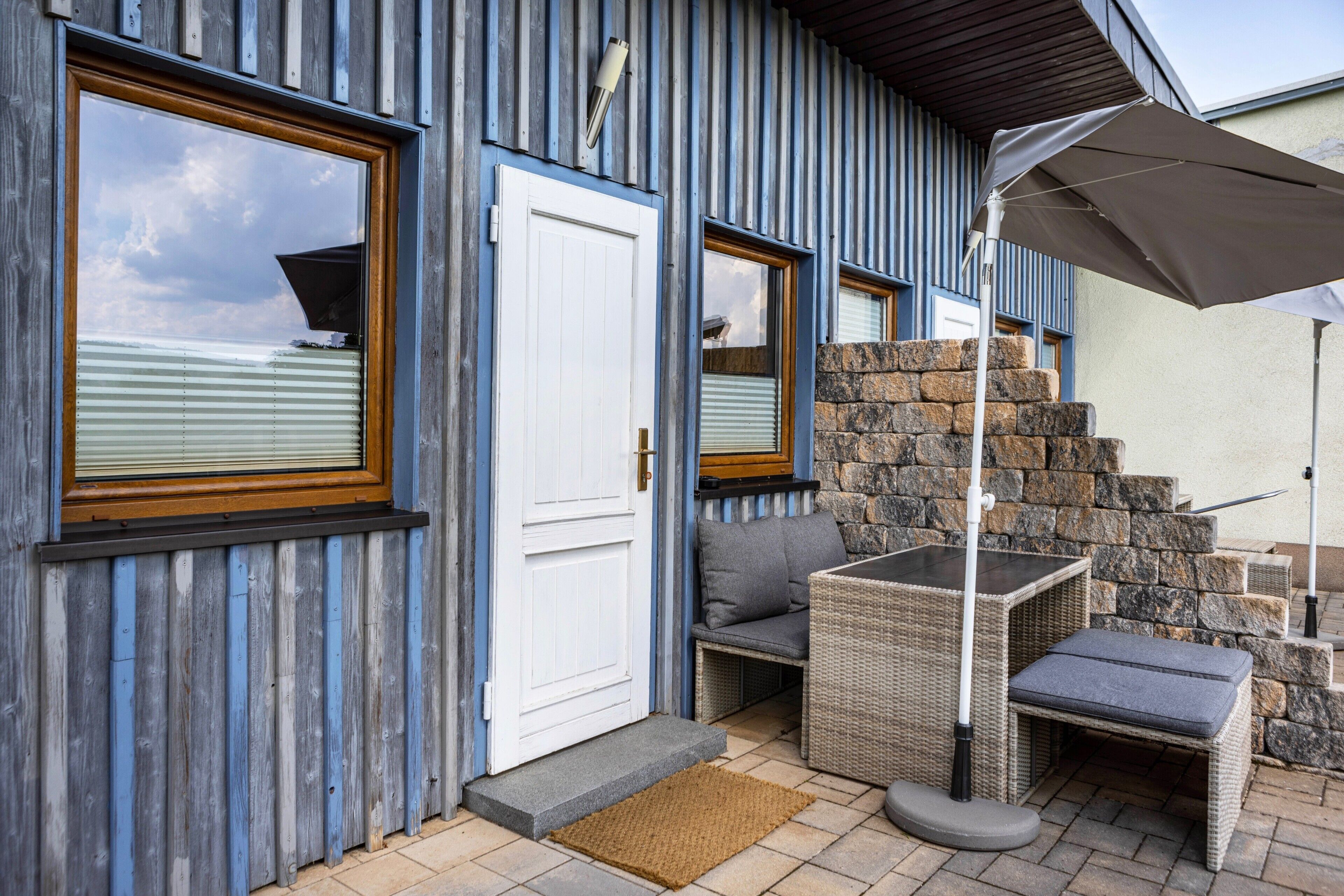 Une façade en bois avec une porte blanche, de grandes fenêtres, un escalier en pierre et du mobilier d’extérieur.