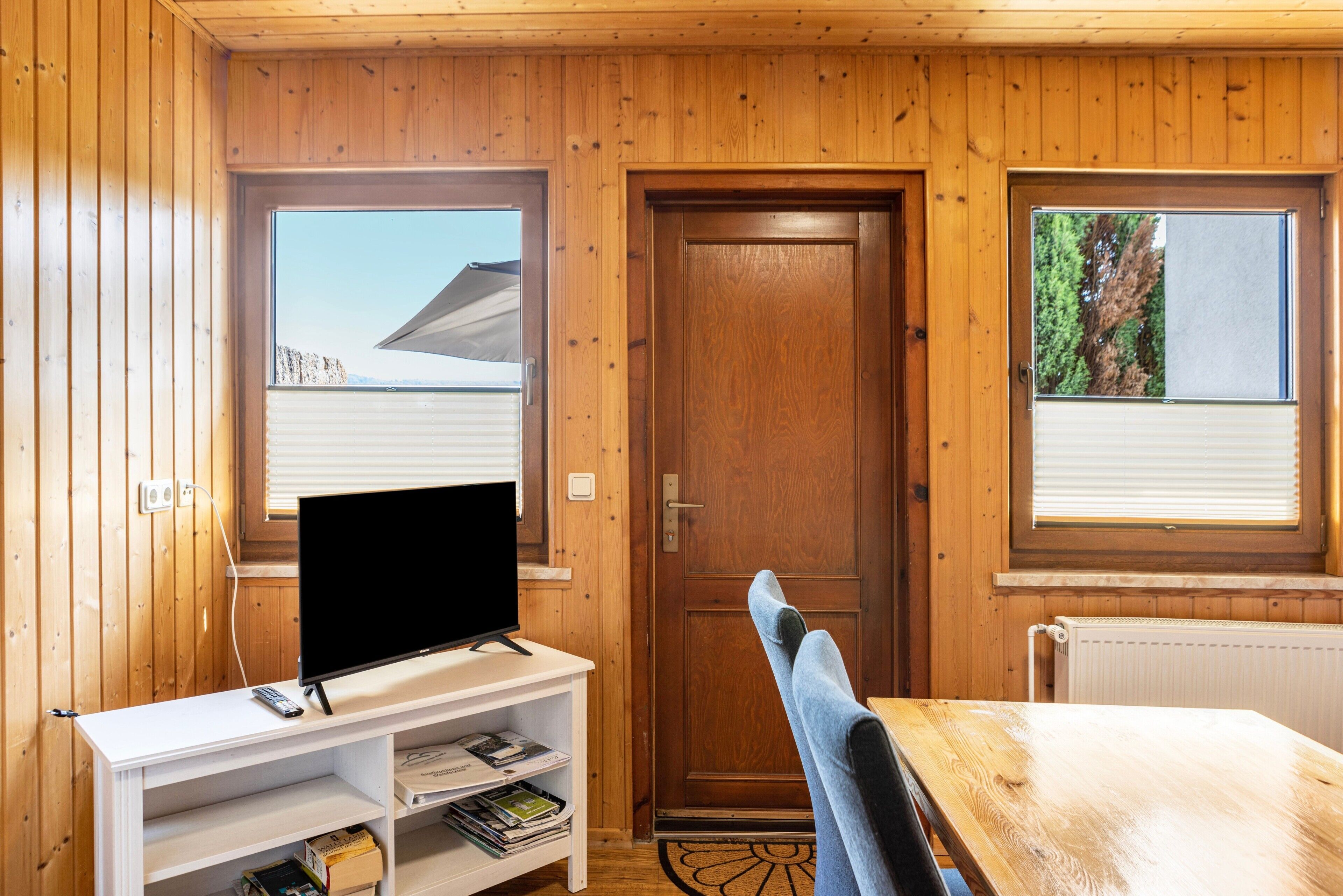 Ein Zimmer mit Holzwänden, einem Fernsehgerät auf einem weißen Schrank, einer Tür und zwei Fenstern.