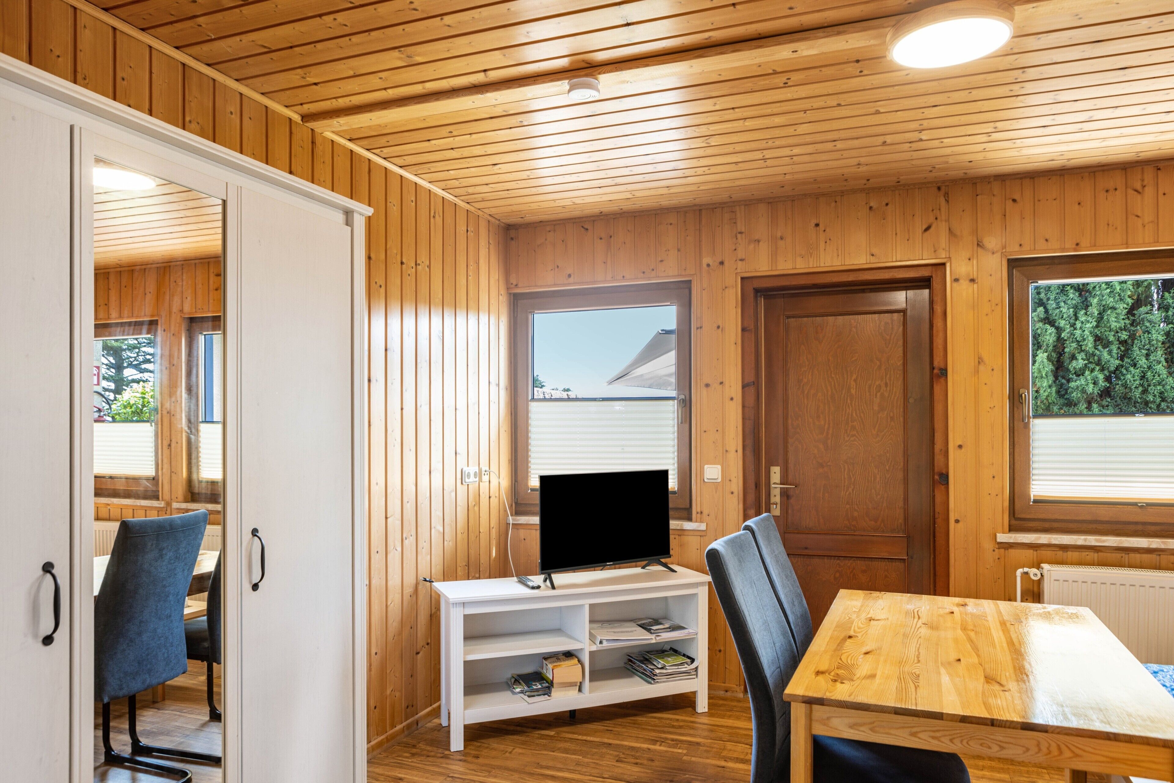 Ein Zimmer mit Holzwänden, einem Fernseher auf einem weißen Standfuß, einem Tisch und Stühlen.