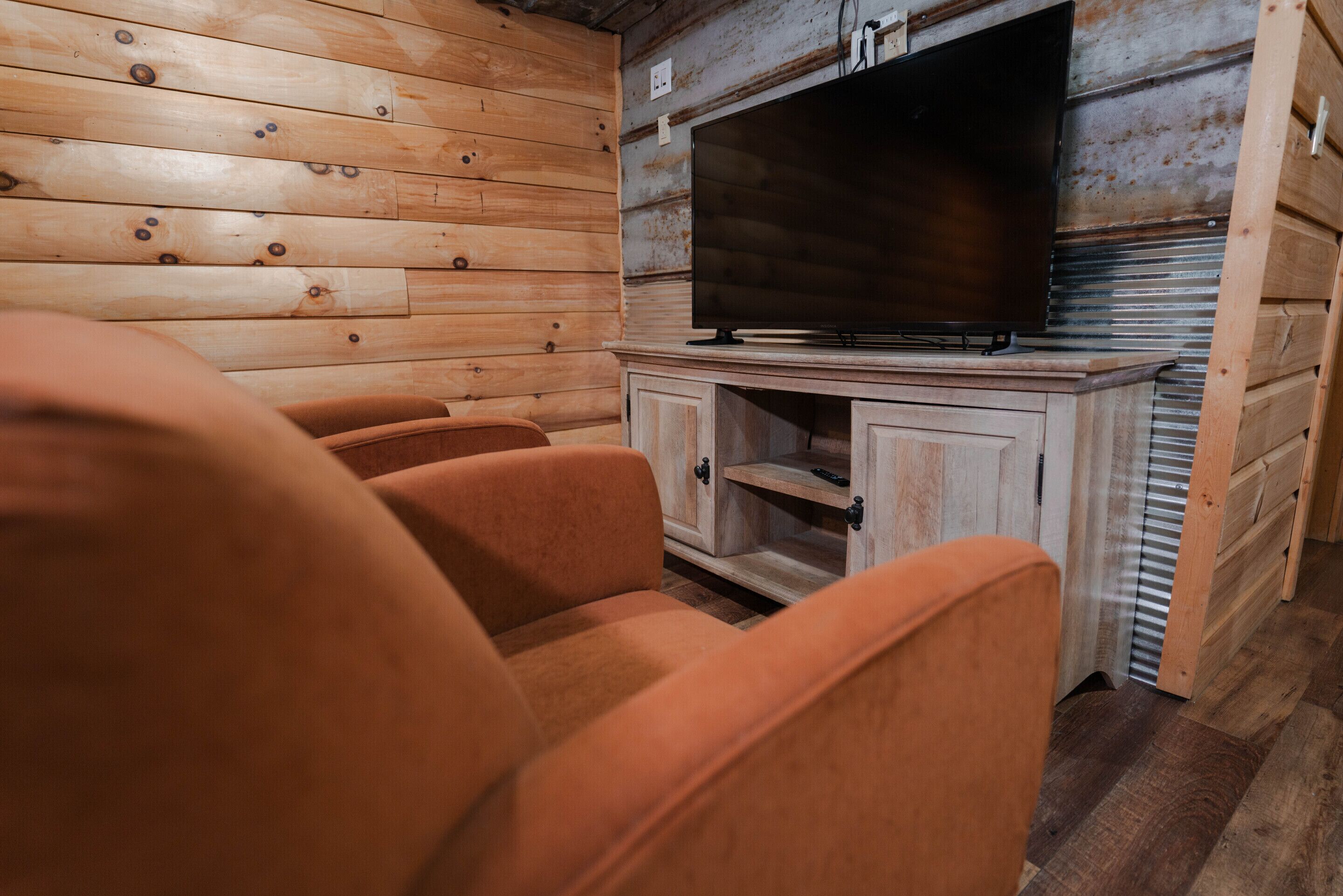 A wooden cabin interior with a television on a cabinet, wooden walls, and orange upholstered chairs.