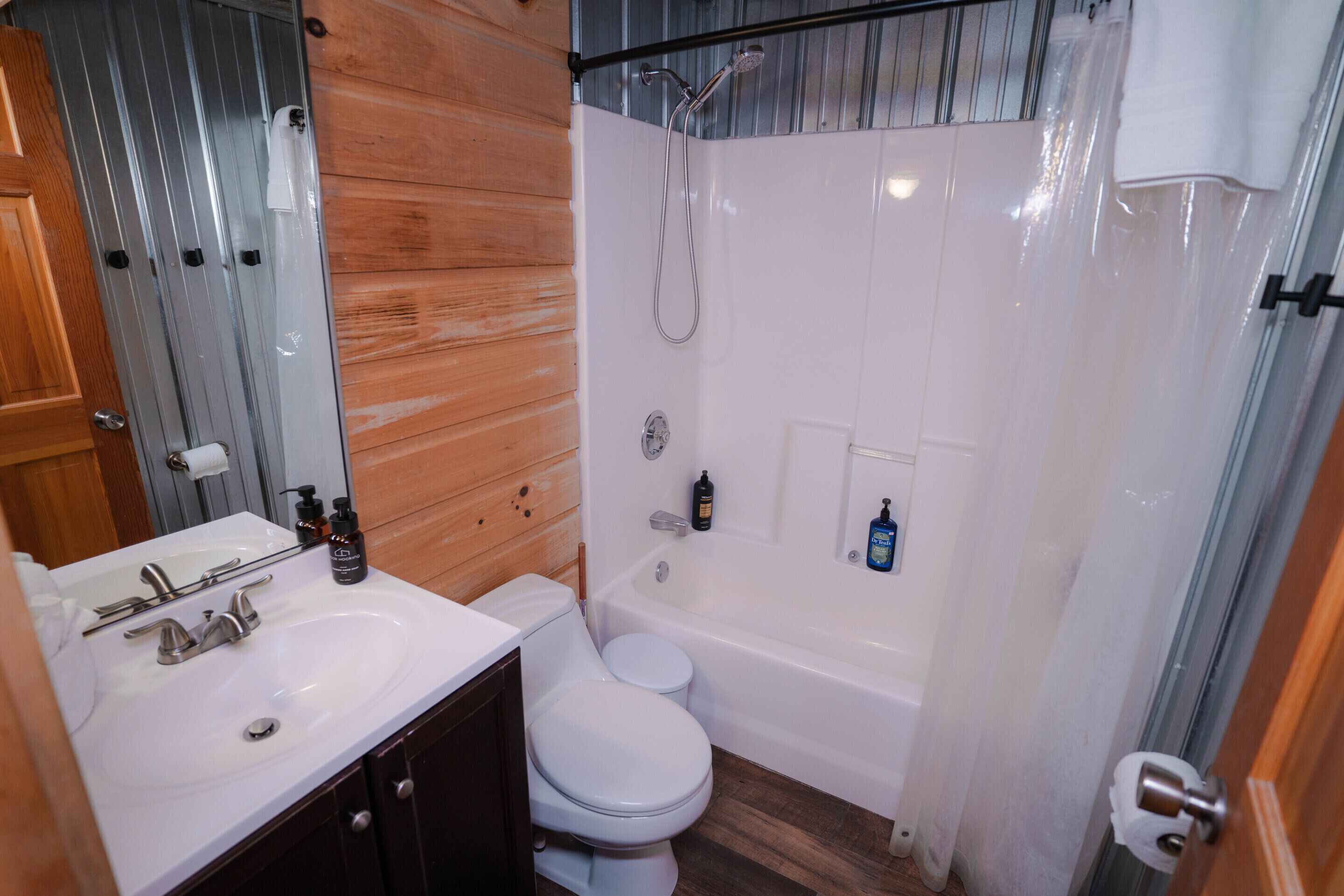 Een badkamer met een wit wastafelmeubel, toilet en badkuip, houten lambrisering aan één wand, en een douchegordijn.