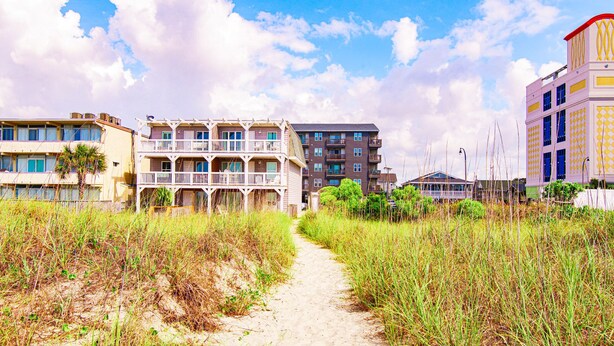 A beachfront hotel with multiple balconies, a sandy path, and surrounding vegetation. - Cherry Grove Condo Steps to Beach (North Myrtle Beach)
