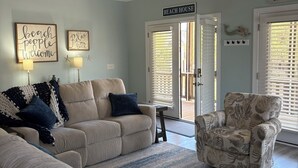 Uma sala de estar com ventilador de teto, um sofá, uma poltrona e uma placa com a inscrição 'BEACH HOUSE'.