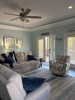 A living room with a ceiling fan, a couch, a chair, and a sign reading 'BEACH HOUSE'. - The Lazy Turtle (Oak Island)