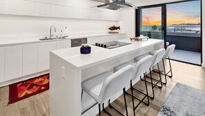 A modern kitchen with white cabinetry, a central island with bar stools, a large window with a view of a sunset, and a red patterned rug. - Bellingham Waterfront Penthouse • Wraparound Balcony, Downtown & Near WWU (Bellingham)