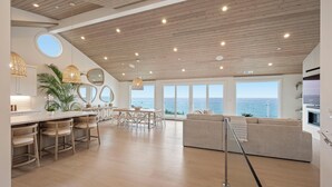 Ett rymligt vardagsrum med ett träpanelat tak, stora fönster med utsikt över havet, en matplats med flätade stolar och ett modernt kök.