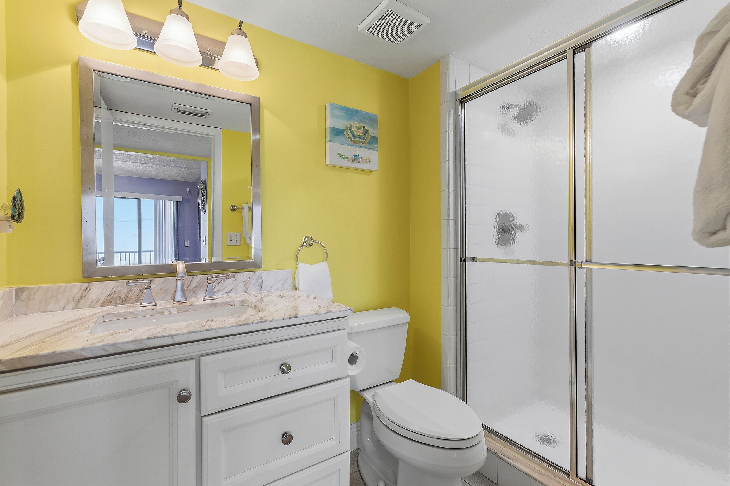A bathroom with a yellow wall, a white toilet, a glass shower enclosure, and a marble countertop.