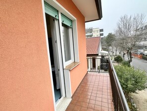 Ein Balkon mit Fliesenboden, ein Fenster mit Vorhang und Blick auf eine Straße mit Gebäuden und Bäumen.