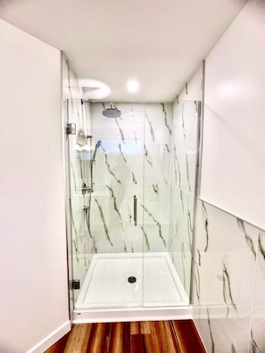 A modern bathroom with a glass-enclosed shower, marble walls, and a wooden floor.