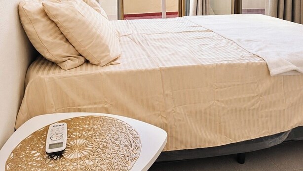 Quarto de hotel com cama, mesa com controle remoto e vista para edifícios através de janelas amplas.