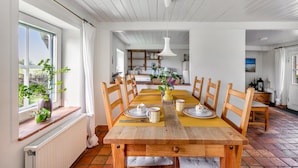 Une salle à manger avec une table en bois dressée pour quatre personnes, des chaises, un luminaire suspendu et une vue sur la cuisine.