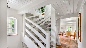 Un escalier blanc menant à un deuxième étage, une fenêtre avec des rideaux et une pièce dotée d’un sol carrelé et d’une chaise.