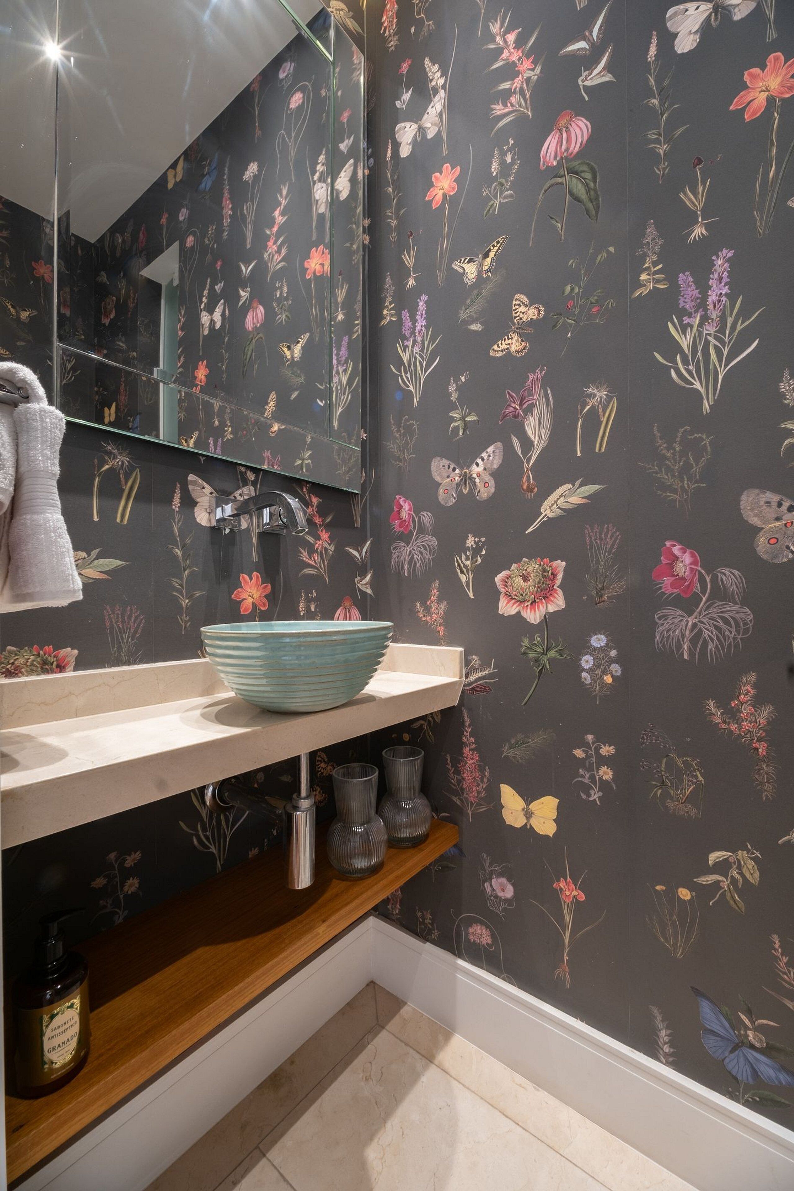 A bathroom with a floral wallpaper, a sink with a blue bowl, a mirror, and a shelf with toiletries.