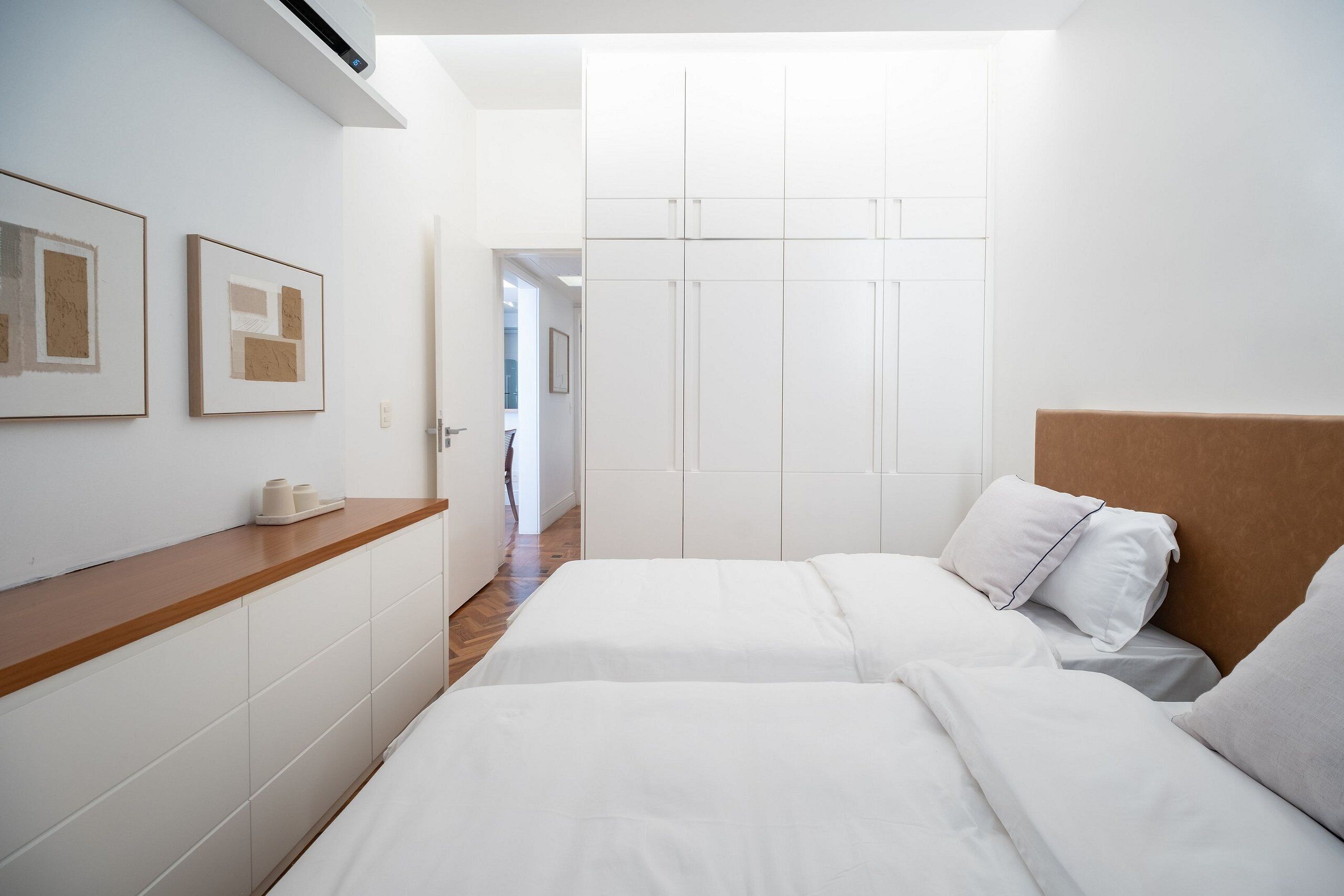 A bedroom with two beds, a wooden bedside table, a white wardrobe, and framed artwork on the wall.