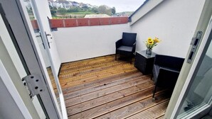 A balcony with a wooden floor, a chair, a small table with a vase of flowers, and a view of houses in the background. - Apartment Sleeps 4 With Balcony and Parking (Kingsbridge)