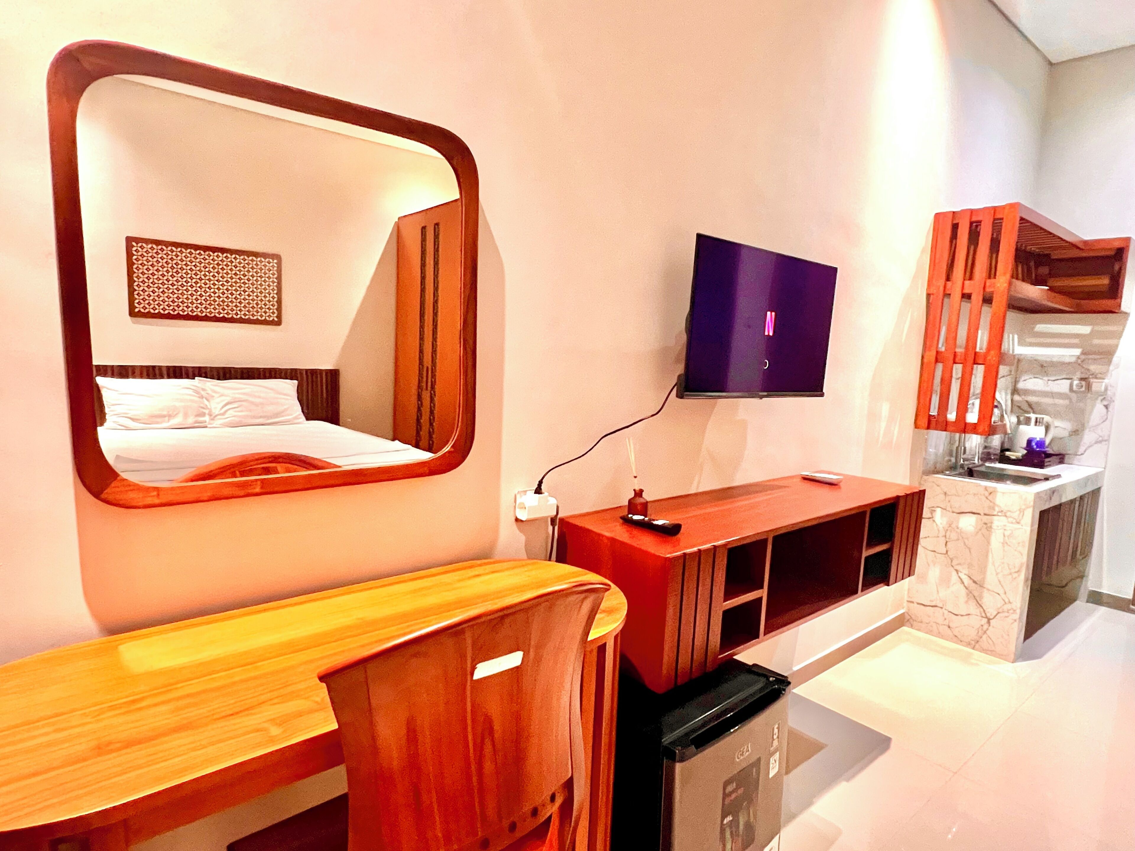Una habitación de hotel moderna con un espejo grande, un televisor montado en la pared, un escritorio de madera y una zona de cocina integrada.