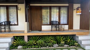A modern building with a covered porch, two chairs, a decorative wall piece, and a garden area with plants and stepping stones. - Diafragma Livin (Nusa Dua)