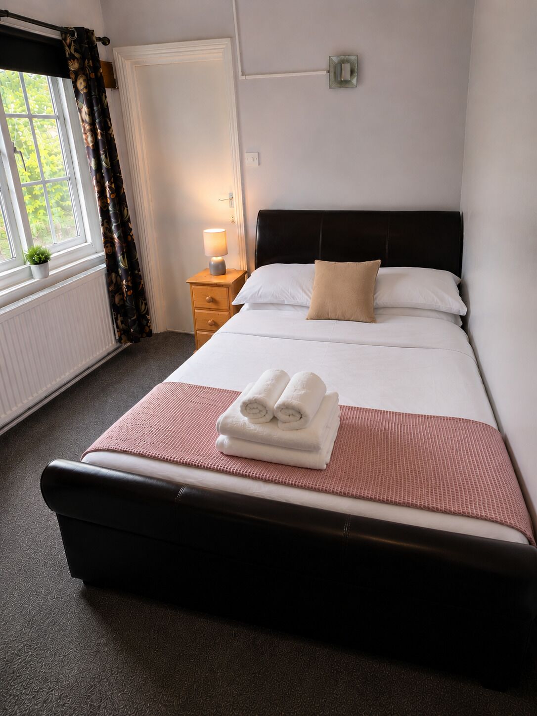 A neatly made bed with white linens and a pink blanket, a bedside table with a lamp, a window with curtains, and a radiator.