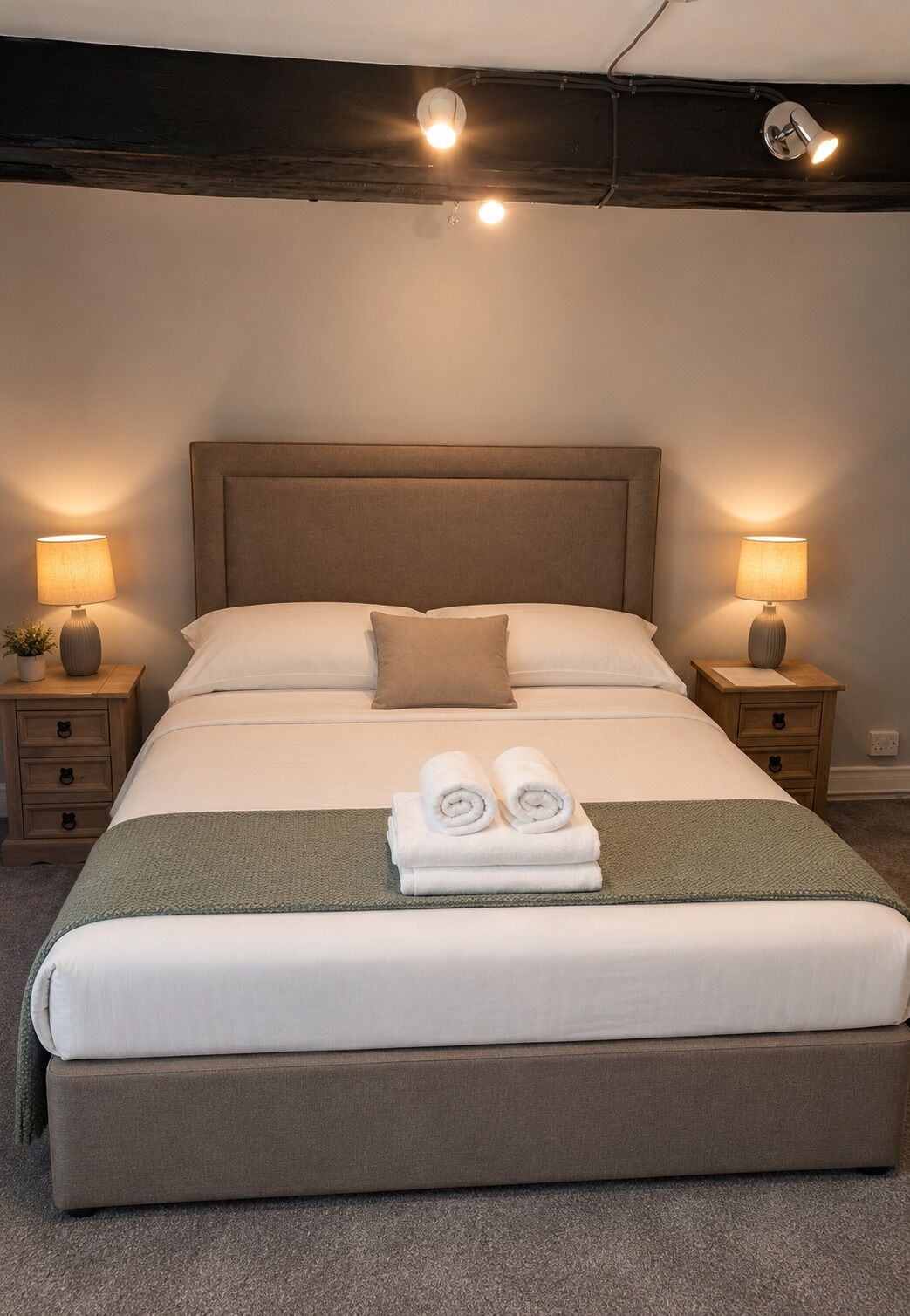 Ein ordentlich gemachtes Bett mit Kopfteil, zwei Nachttische mit Lampen und ein Paar zusammengerollte Handtücher auf dem Bett.