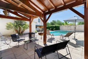 Un patio exterior con piscina, pérgola de madera y sillones de descanso.