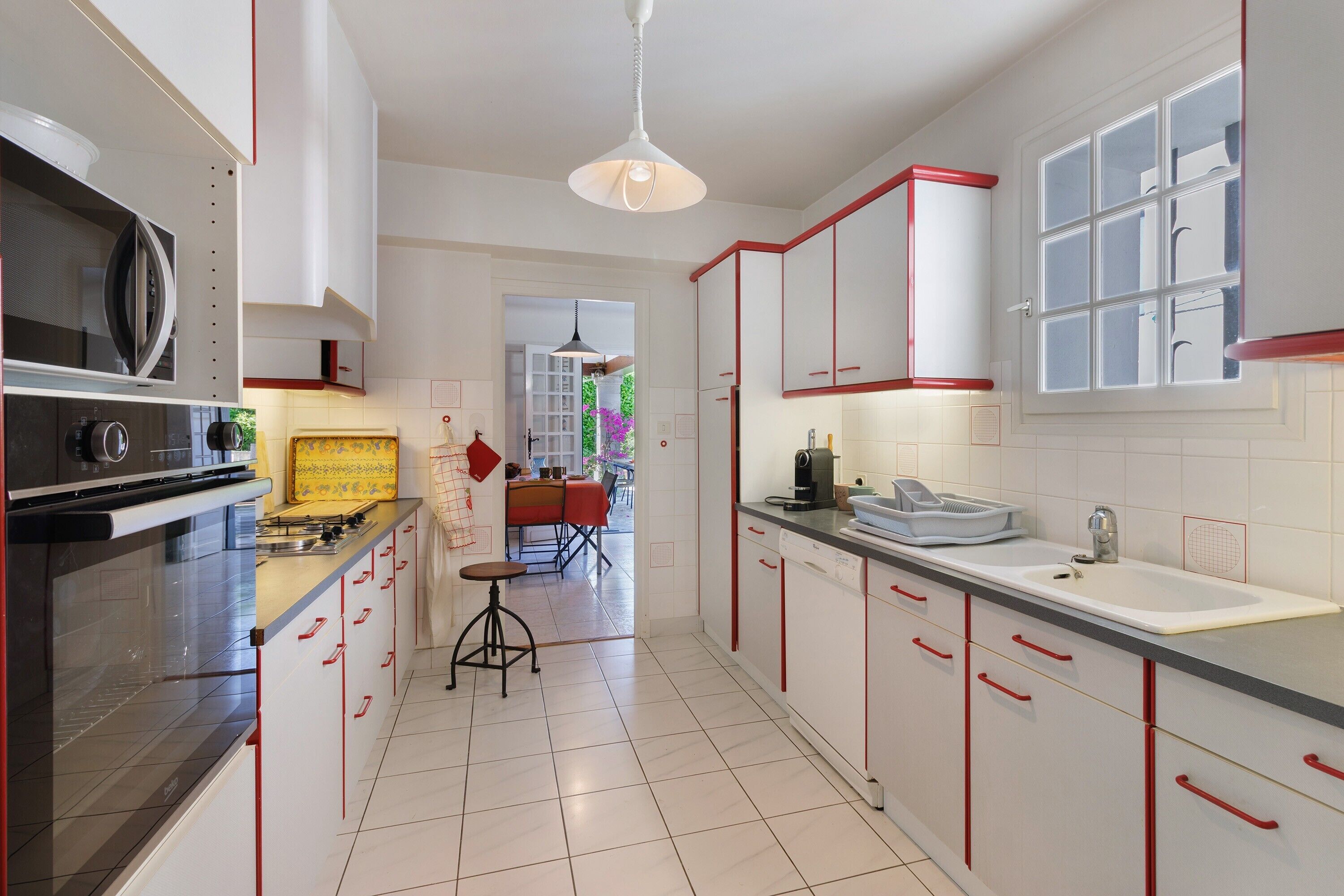 A modern kitchen with white cabinets, red handles, a built-in oven, and a dining area with a red table and chairs.