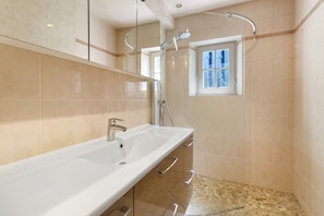 Un baño con un lavamanos blanco, cabina de ducha con mampara de vidrio y una ventana.