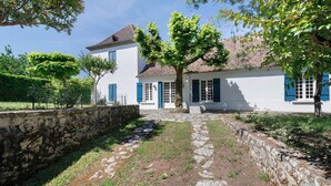 A stone pathway leading to a white building with blue shutters, surrounded by greenery and trees. - Le Tsilaosa – Charm and Relaxation in the Heart of Périgord (Prigonrieux)