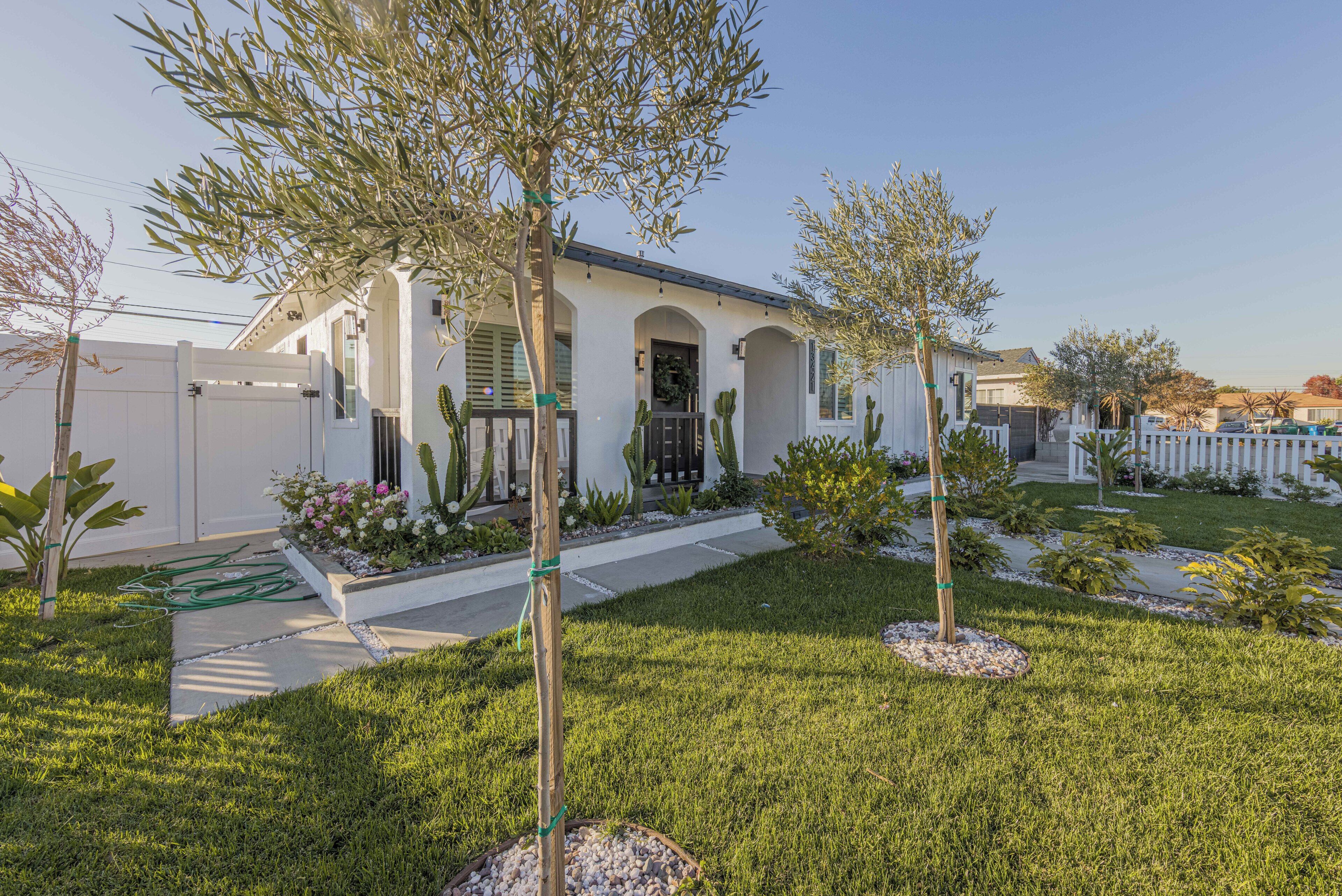 A single-story house with a white exterior, a front porch, and a well-maintained lawn with small trees and shrubs.