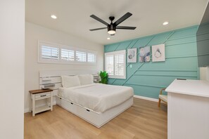 Un dormitorio con cama, mesitas de noche, escritorio, ventilador de techo y cuadros en la pared.