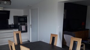 En matplats med ett rektangulärt bord och fyra trästolar, ett kök med moderna vitvaror och ett vardagsrum med mörka färgtoner.