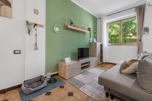Sala de estar con sofá, televisor, mueble de madera para entretenimiento, cama para mascotas y ventana con vista a árboles.