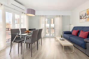 Ein modernes Wohnzimmer mit einem Glastisch, grauen Stühlen, einem blauen Sofa mit roten Kissen und einem großen Fenster mit Vorhängen.