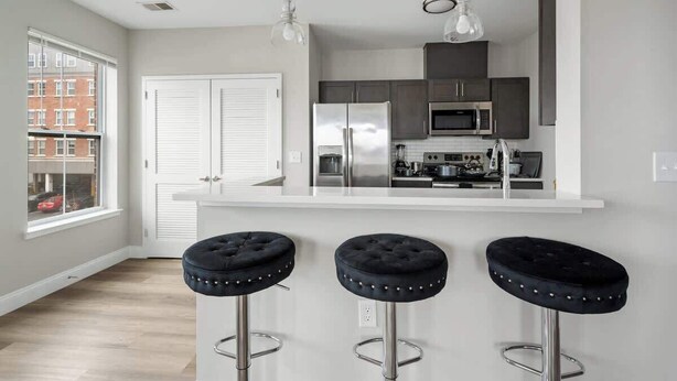 A modern kitchen with a white island, stainless steel appliances, and three black bar stools. - LuxBay Place 1BD/5 Min to Rutgers Newark/Pkg Incl (Newark)