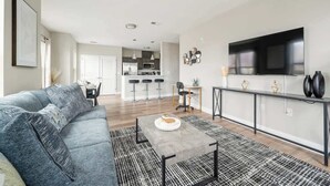 A modern living room with a grey sofa, a coffee table, and a TV. There is a kitchen in the background with bar stools. - LuxBay Place 1BD/5 Min to Rutgers Newark/Pkg Incl (Newark)