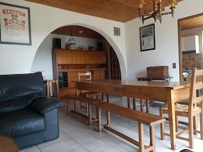 Ein Essbereich mit Holztisch und Bänken, einem schwarzen Ledersofa und einer Küche im Hintergrund.