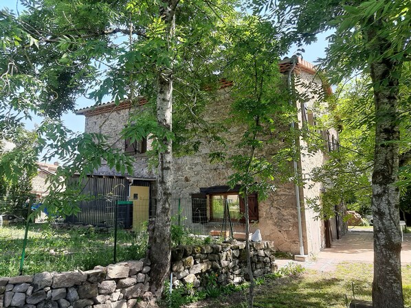 Ein Gebäude aus Stein mit rustikaler Fassade, eine Steinmauer und ein Gartenbereich mit Bäumen und Pflanzen.