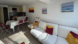 A modern living room with a white sofa, red and yellow cushions, a dining area with a white table and red chairs, and a kitchen in the background. - Appartement Camargue Village à deux pas de la plage du Boucanet (Le Grau-du-Roi)