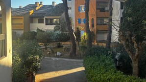 A courtyard with a paved path, surrounded by trees and shrubs, leading to a building with a tiled roof and multiple windows. - Appartement Camargue Village à deux pas de la plage du Boucanet (Le Grau-du-Roi)
