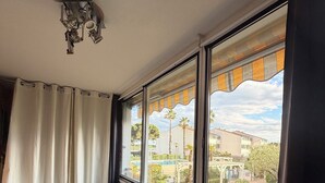 A dining area with a table set for two, a chair, and a view of a courtyard with palm trees and a building. - Terrasses du Lac – vue lac piscine clim parking terrasse (Le Grau-du-Roi)