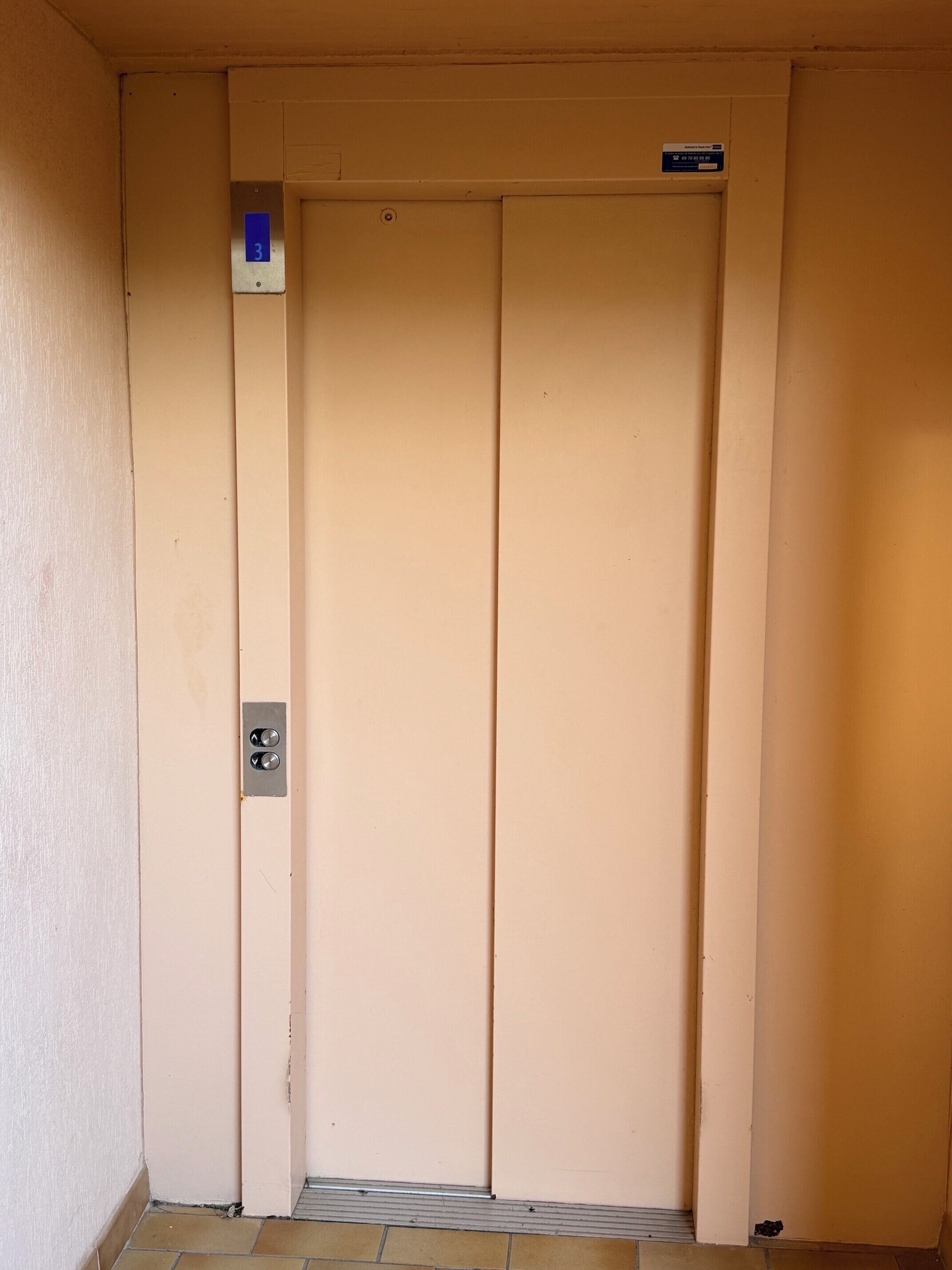 Un ascenseur de couleur beige, doté d’un panneau de commande et d’une enseigne bleue au-dessus.