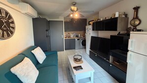 A compact living space with a sofa, a coffee table, a TV, a kitchen area, and a wall clock. - Terrasses du Lac – vue lac piscine clim parking terrasse (Le Grau-du-Roi)